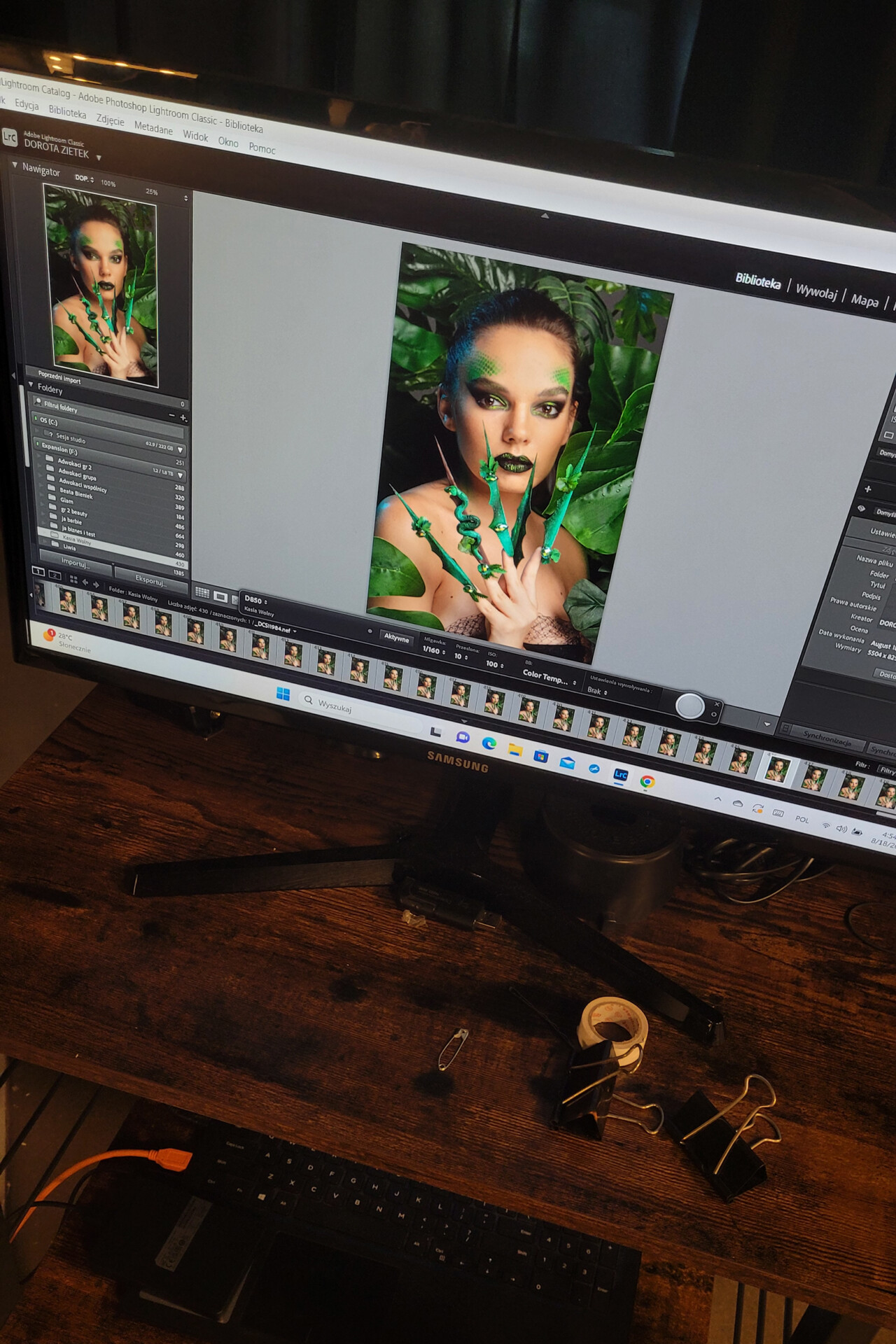 Zdjęcie osoby z wyrazistym makijażem i stylizacją paznokci, prezentującej dłonie w zielonych akrylowych paznokciach, otoczona zielonymi roślinami na ekranie komputera. W tle widoczny stół z narzędziami do stylizacji i biurowymi przyborami