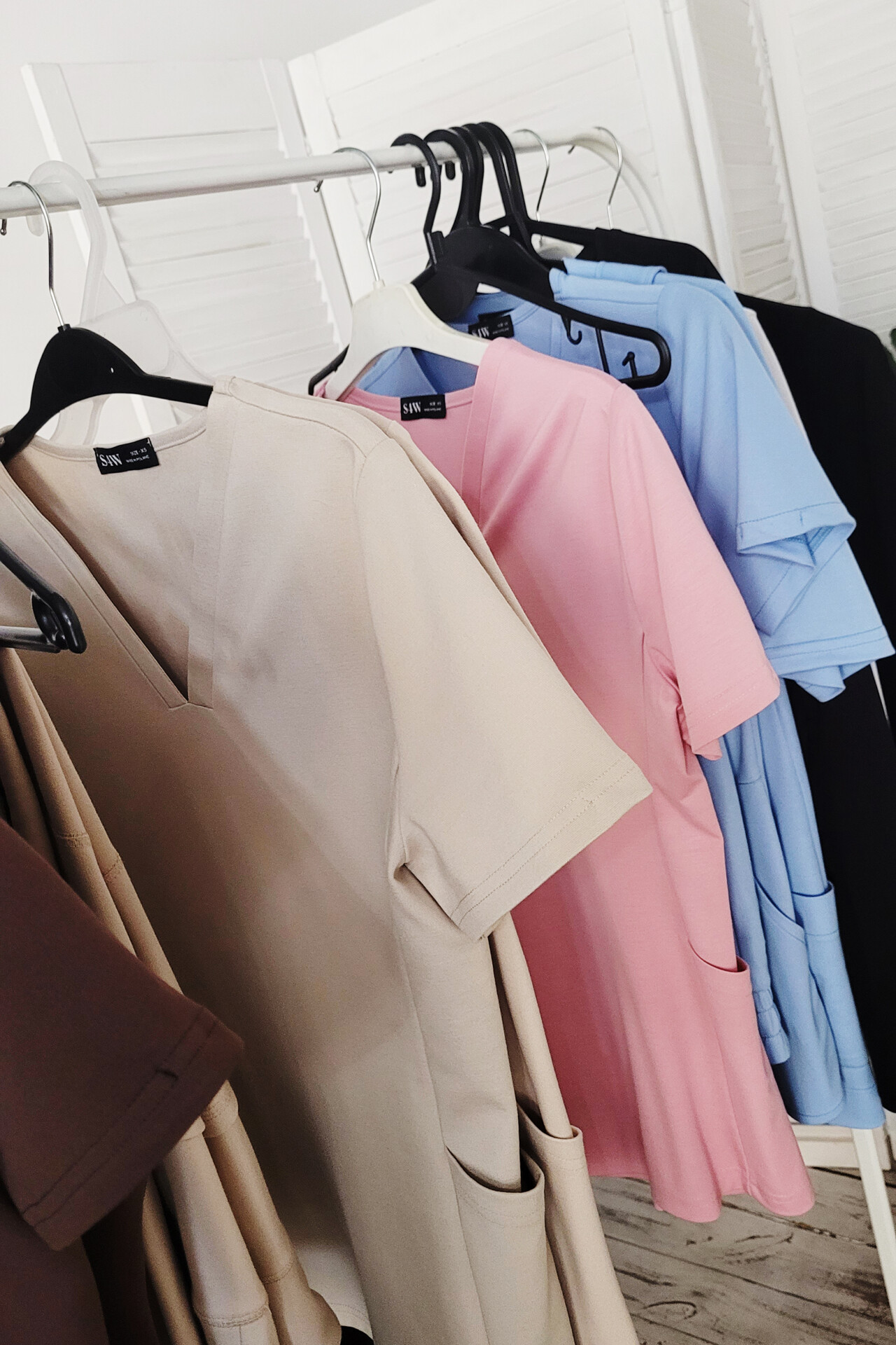 Na wieszaku widać kilka kolorowych bluzek medycznych, w odcieniach beżowym, różowym, niebieskim i czarnym. Każda bluzka ma krótki rękaw oraz kieszenie po bokach