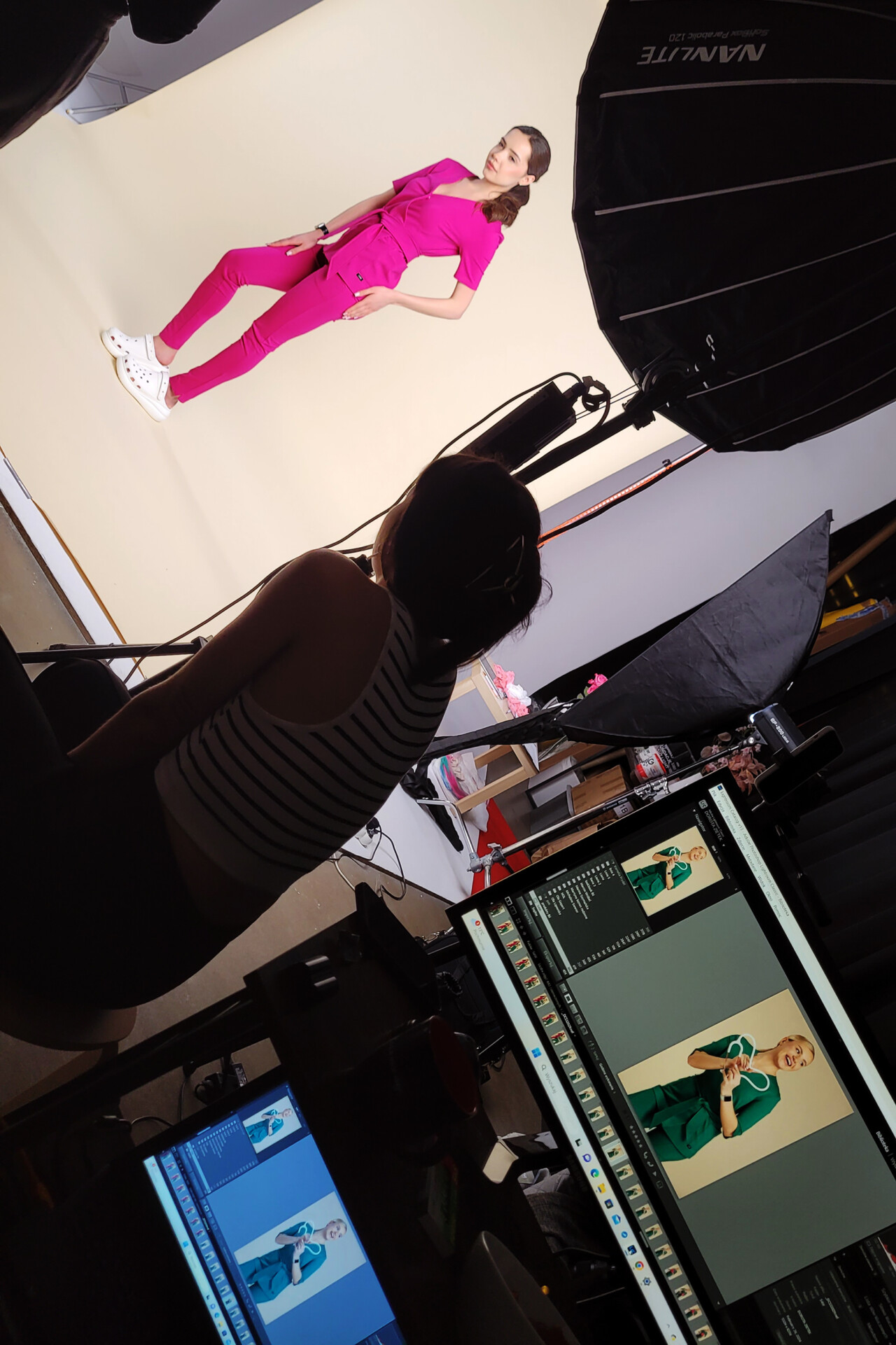 Modelka w różowym ubraniu stoi na tle jasnej ściany, podczas sesji zdjęciowej. W tle widać sprzęt fotograficzny i dwa monitory z podglądem zdjęć