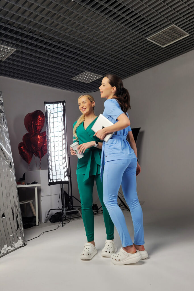 Dwie kobiety w kolorowych strojach medycznych: jedna w zielonym, druga w niebieskim, pozują razem w studio zdjęciowym, w tle balony w kształcie serca