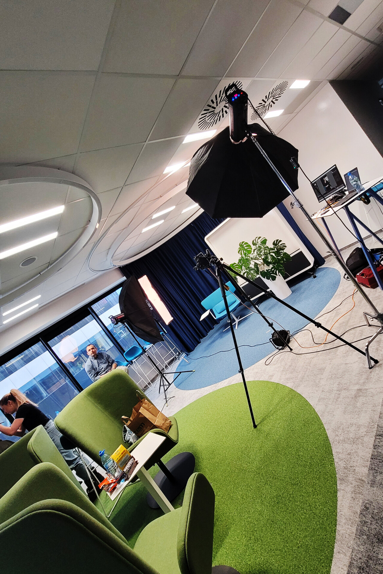 W biurze z zielonym dywanem i nowoczesnymi meblami, stoją dwa statywy z oświetleniem. W tle widoczna jest osoba siedząca w fotelu. Na stole znajdują się laptop i inne akcesoria
