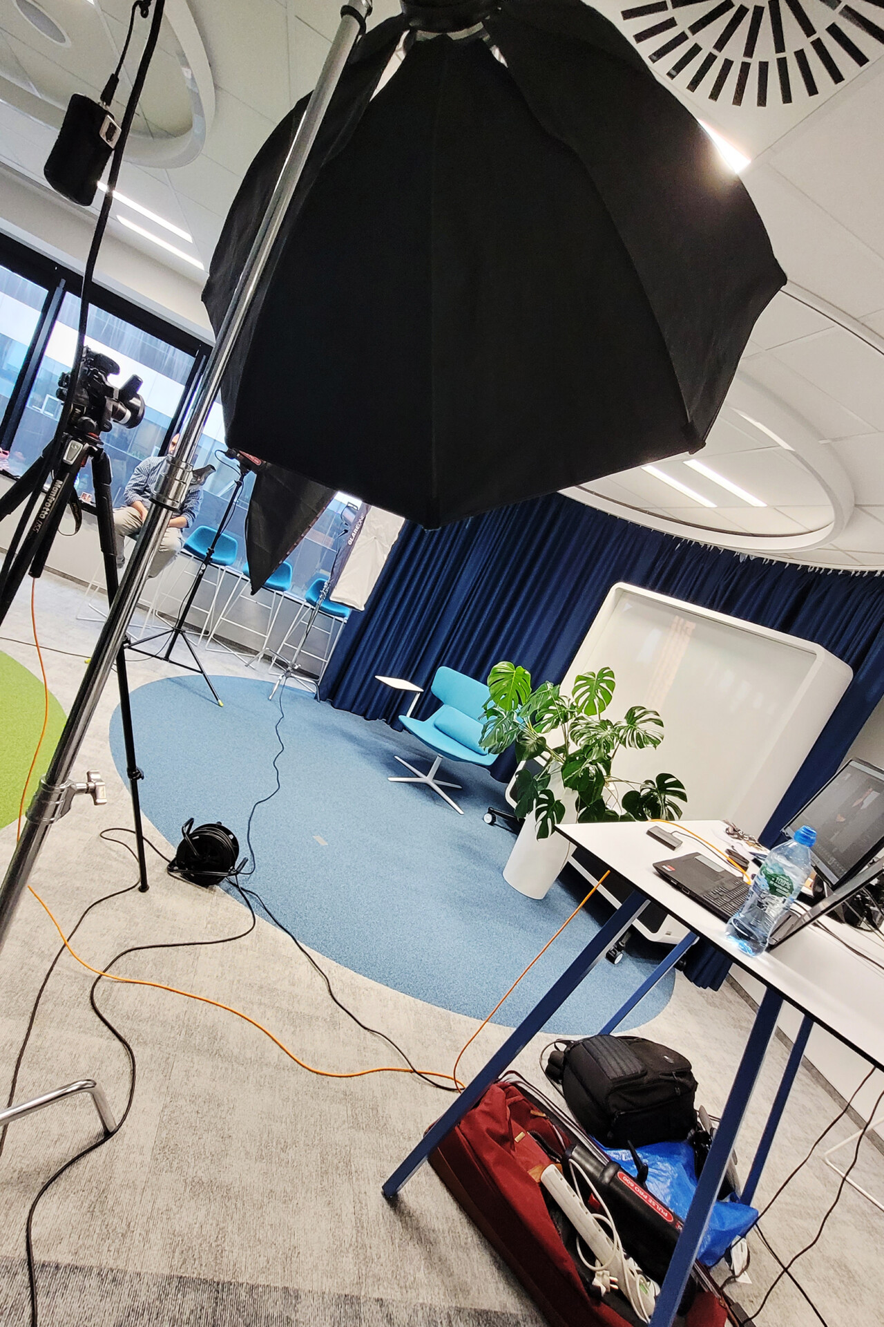 W biurze znajduje się sprzęt do nagrywania, w tym statyw z oświetleniem i kamery. W tle widoczna jest roślina oraz niebieskie krzesło przy stoliku. Podłoże jest w jasnych kolorach, a w pomieszczeniu panuje jasne oświetlenie