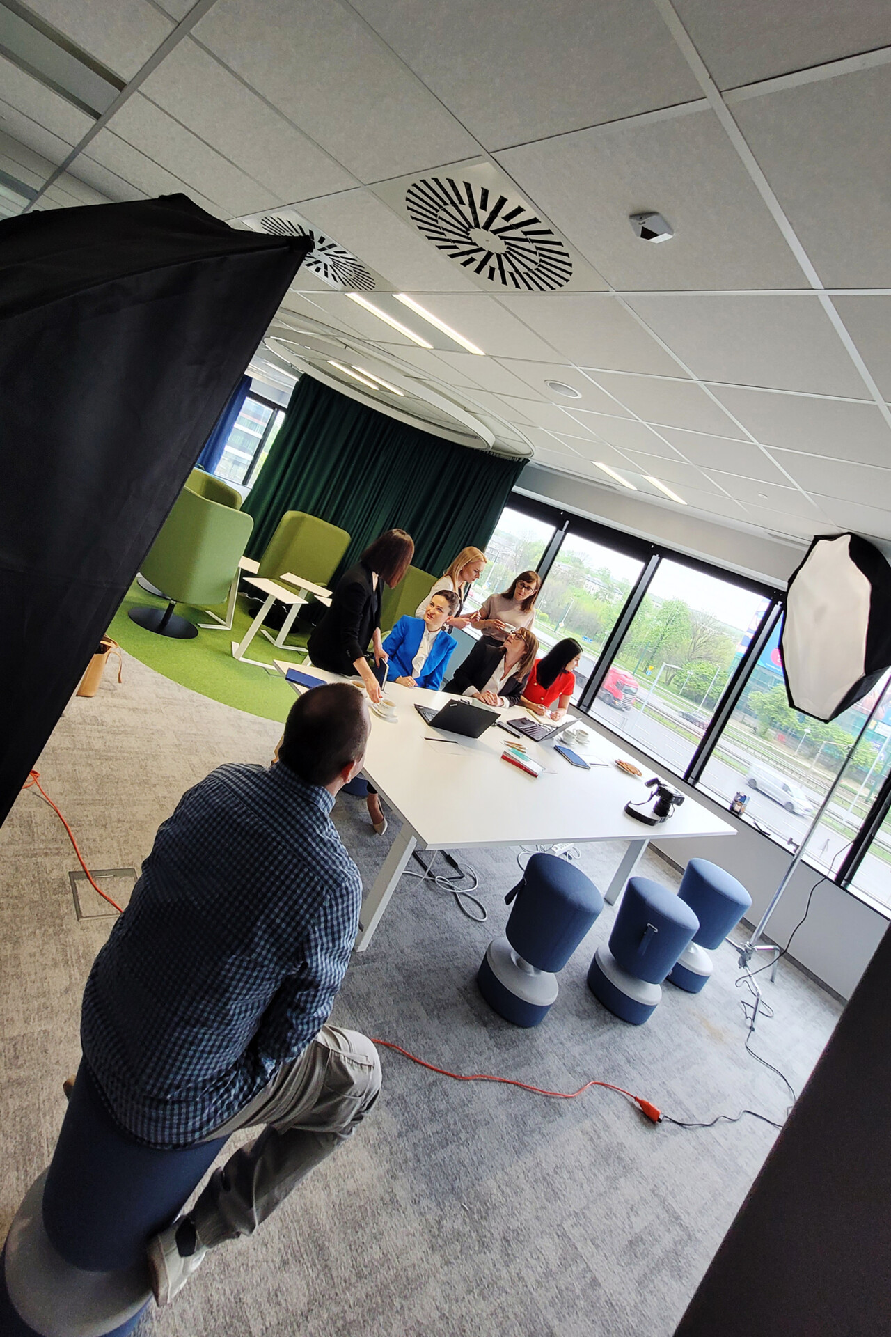 Grupa pracowników TUV Rheinland Polska prowadzi sesję biznesową w nowoczesnym biurze, z dużymi oknami i zielonymi elementami wystroju. Przy stole są laptop i kilka osób, podczas gdy jedna siedzi na stołku z tyłu