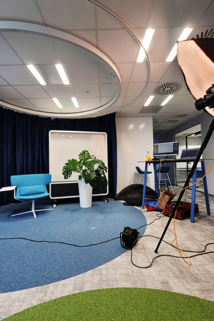 Nowoczesne biuro z niebieskim dywanem i zielonymi akcentami, z białą rośliną w doniczce, niebieskim fotelem i sprzętem fotograficznym w rogu