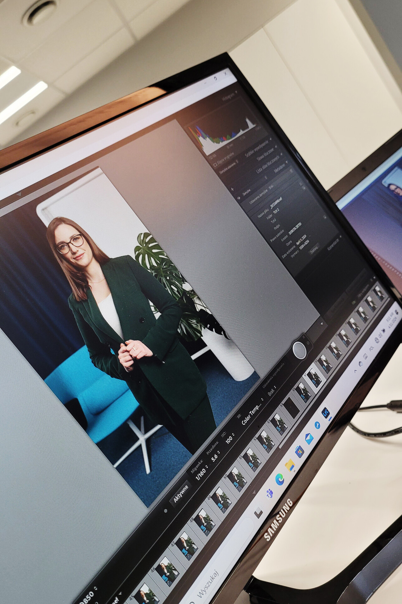 Kobieta w ciemnozielonym żakiecie z okularami stoi wyprostowana w biurze przed rośliną doniczkową, na monitorze widoczna edycja zdjęcia