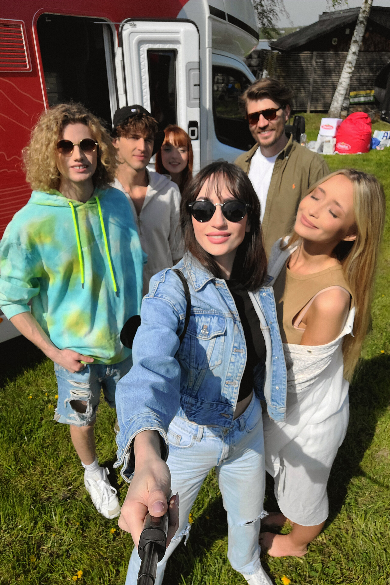 Grupa pięciu osób stoi na świeżym powietrzu, pozując do selfie. Ubrani w kolorowe i luźne stroje, na tle kampera i otaczającej zieleni. Słoneczna pogoda