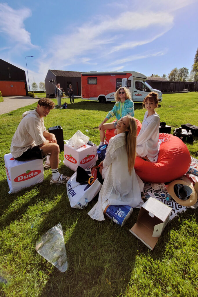 Grupa czterech osób siedzi na trawie w słoneczny dzień. W tle widać samochód dostawczy oraz kilka osób. Obok nich leżą torby i inne przedmioty. Osoby mają różne ubrania i uśmiechają się do kamery