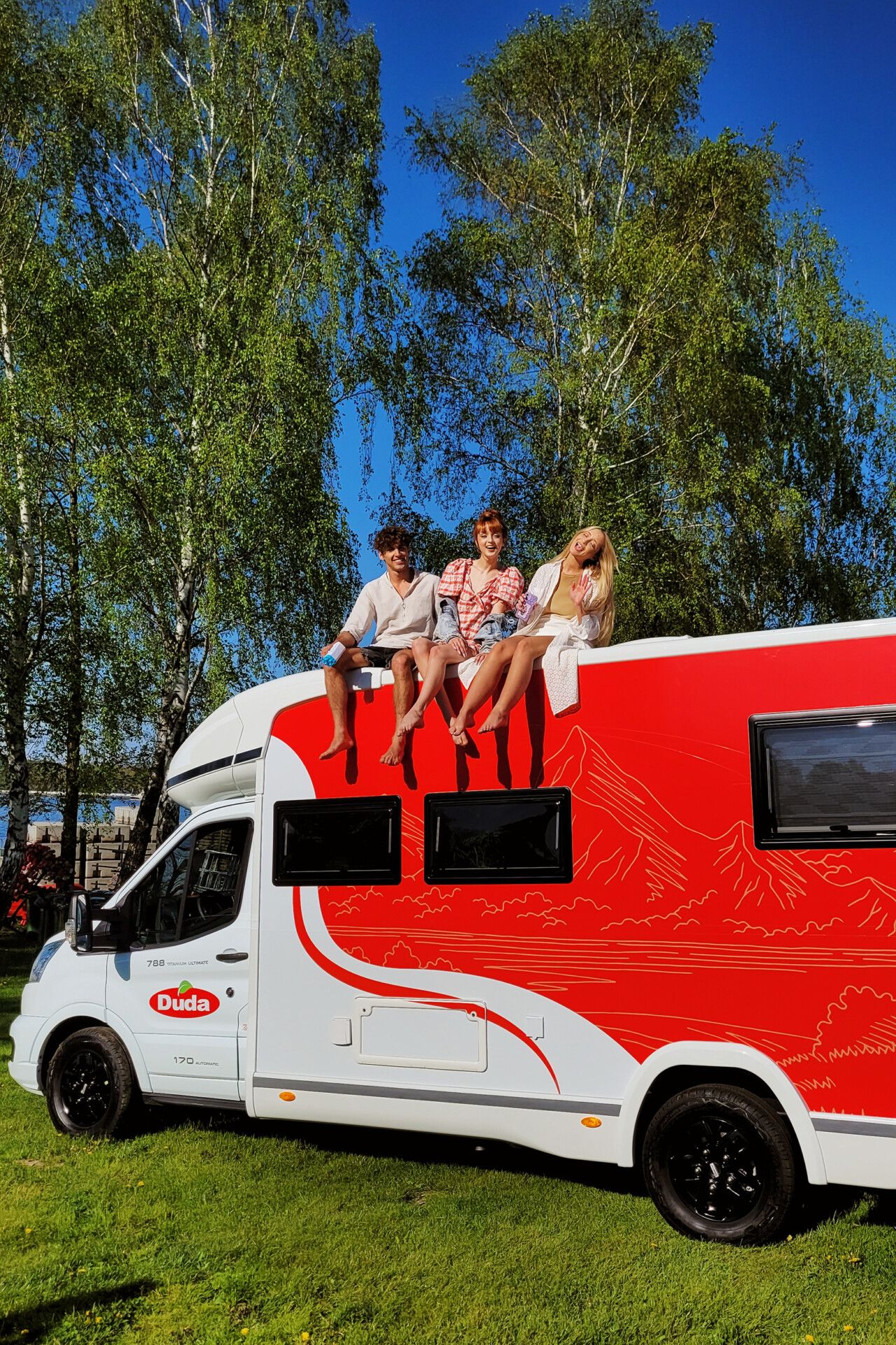 Trzy osoby siedzą na dachu białego kampera z czerwonym wzorem. W tle widać zieloną trawę i drzewa. Dwie osoby mają na sobie jasne ubrania, jedna jest w staniku i szortach. Słońce świeci na bezchmurne niebo