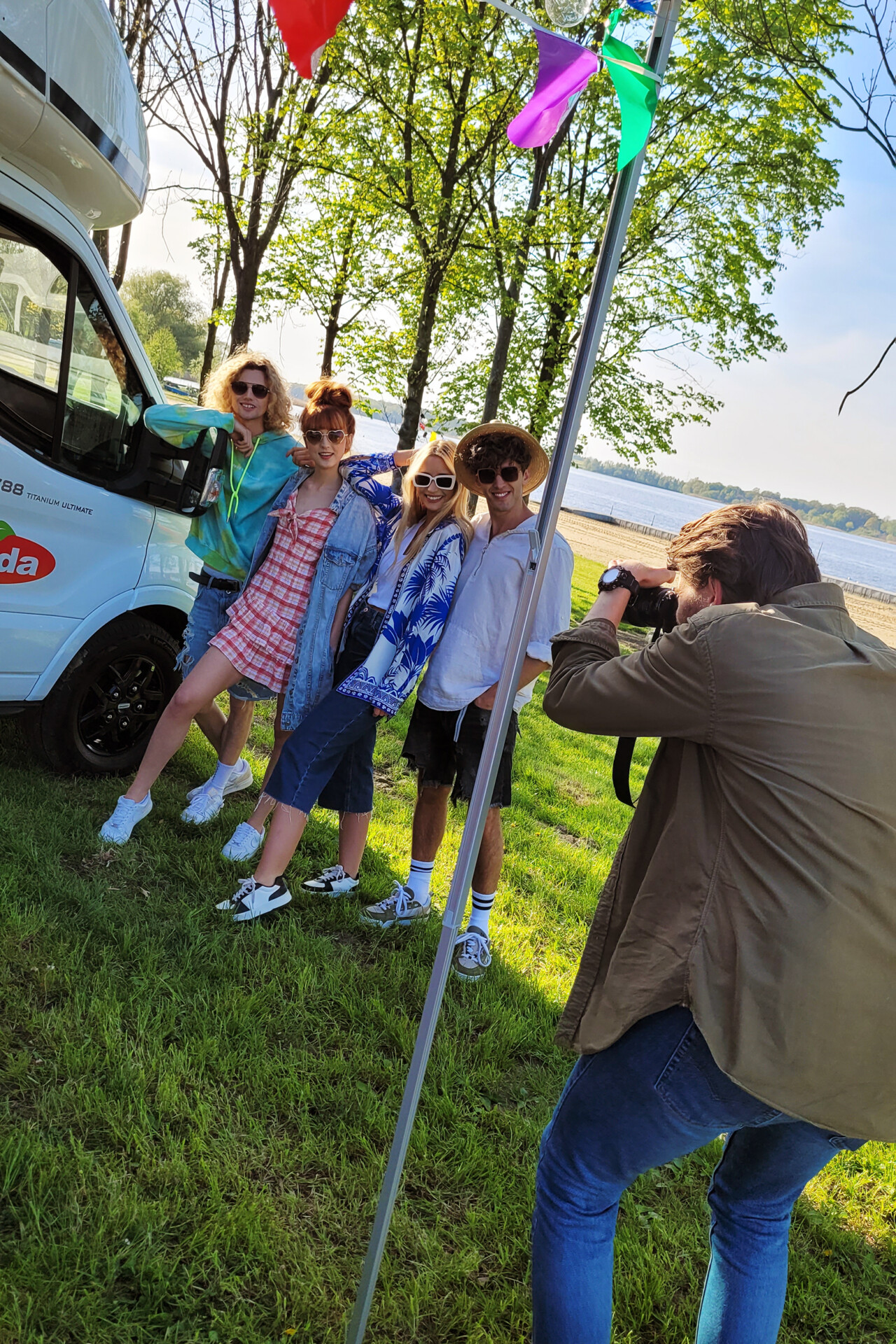 Grupa czterech młodych osób pozująca na trawie w pobliżu samochodu kempingowego. Jedna osoba robi zdjęcie grupie, która uśmiecha się i przyjmuje różne pozy. W tle widać drzewa oraz wodę