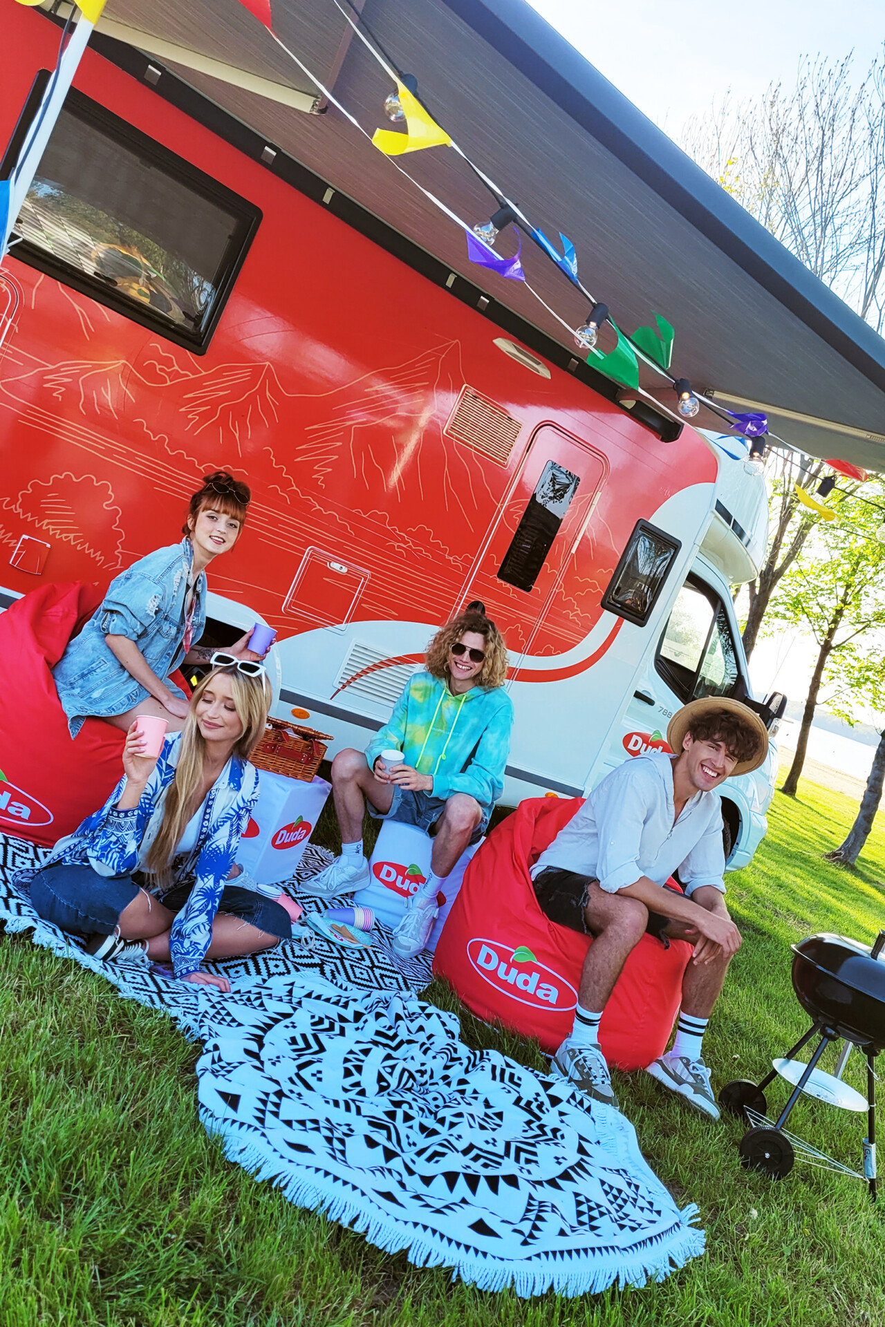 Czworo młodych ludzi w kolorowych ubraniach siedzi na zewnątrz obok przyczepy kempingowej Duda. W tle widać drzewka i flagi, na ziemi leży niebieski dywan. Osoby pozują w luźnej atmosferze przy grillu