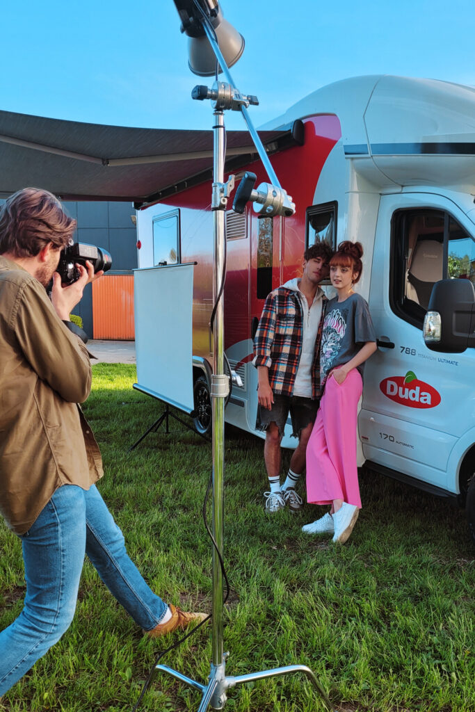 Fotografia przedstawia dwóch modeli pozujących obok białego kampera z logo "Duda". Fotografuje ich osoba trzymająca aparat, trójnożny statyw jest widoczny na pierwszym planie. Na tle widać niebieskie niebo i część zielonego otoczenia. Modelka w różowych spodniach stoi blisko modela, który ją obejmuje