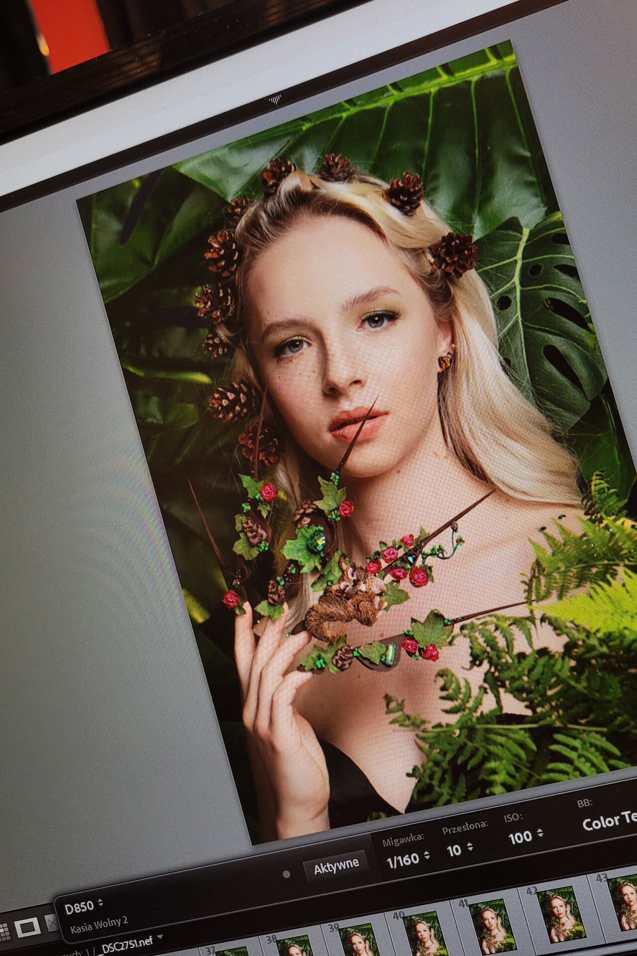 Portret kobiety z długimi blond włosami, trzymającej gałązkę z liśćmi i owocami, otoczonej zielonymi roślinami i szyszkami