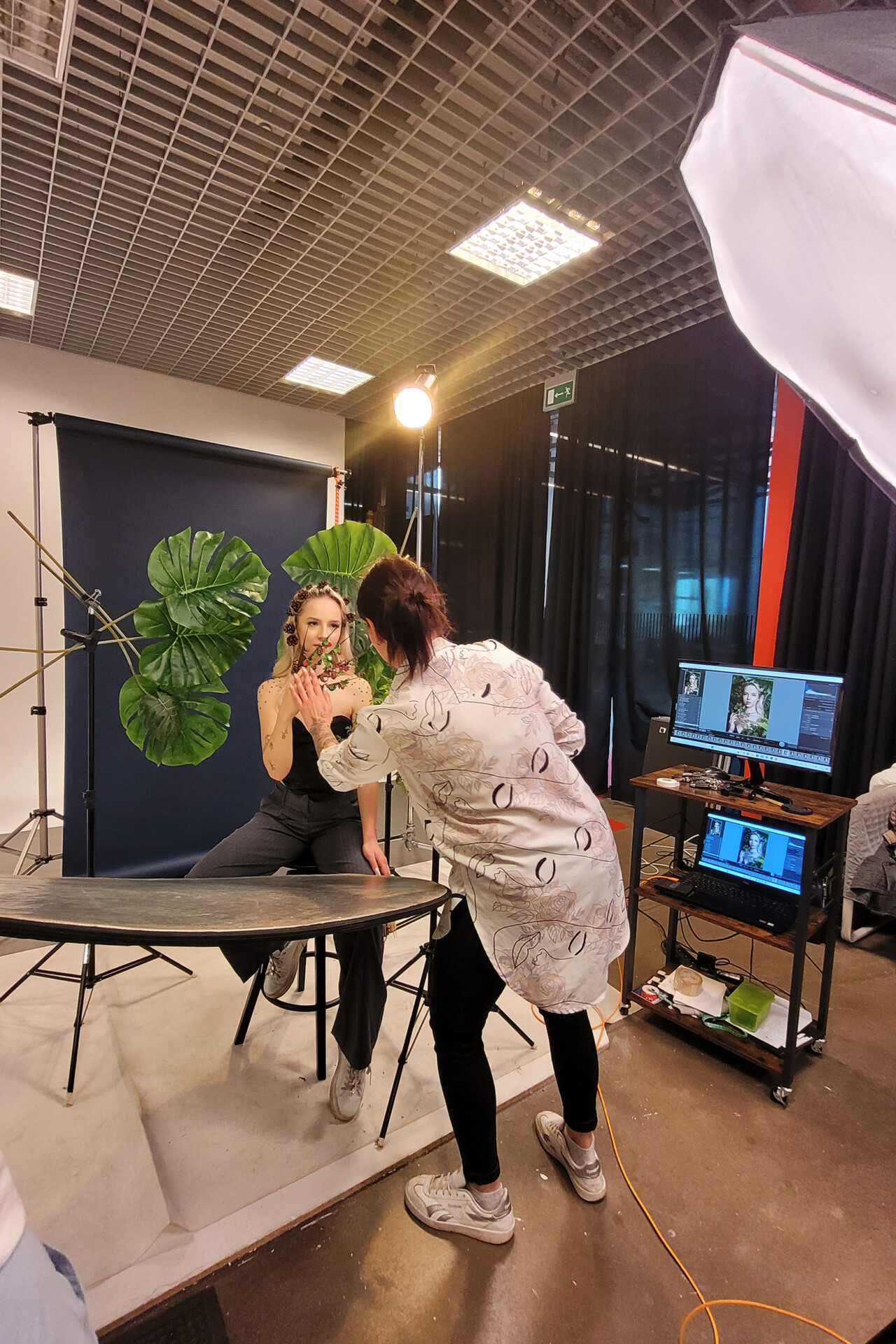 Instruktor stylizacji paznokci siedzi na stołku w studiu, ubrany w szary strój. Osoba obok niego wykonuje manicure, a w tle widoczna jest roślina i oświetlenie studyjne