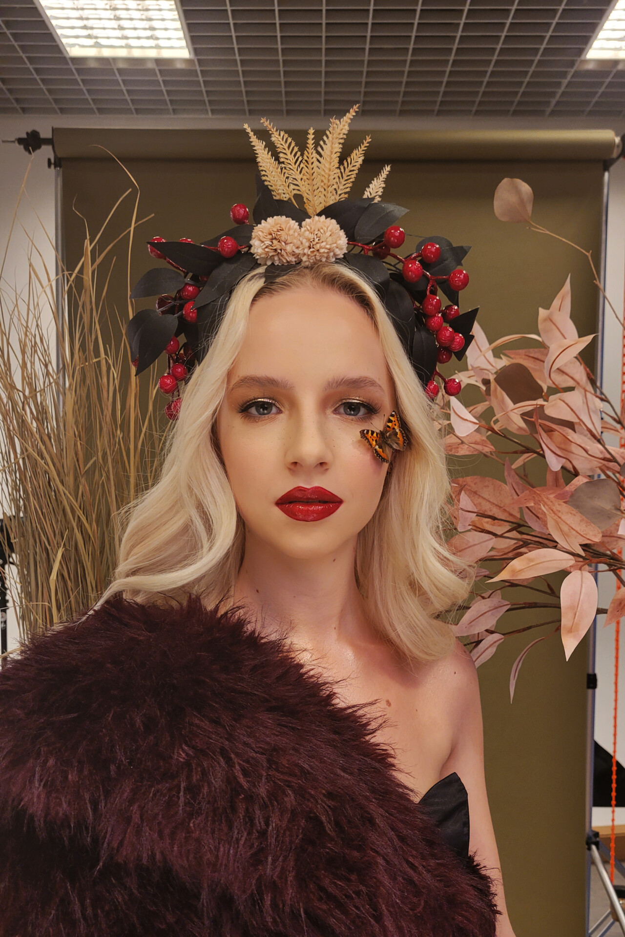 Kobieta z długimi, jasnymi włosami, ubrana w czarną suknię, z makijażem w odcieniach czerwieni i ustami w kolorze czerwonej szminki. Na głowie ma ozdobną opaskę z liśćmi, kwiatami i czerwonymi owocami. Obok niej znajdują się gałęzie roślin w odcieniach beżu i różu. Na twarzy siedzi motyl, dodając artystycznego akcentu