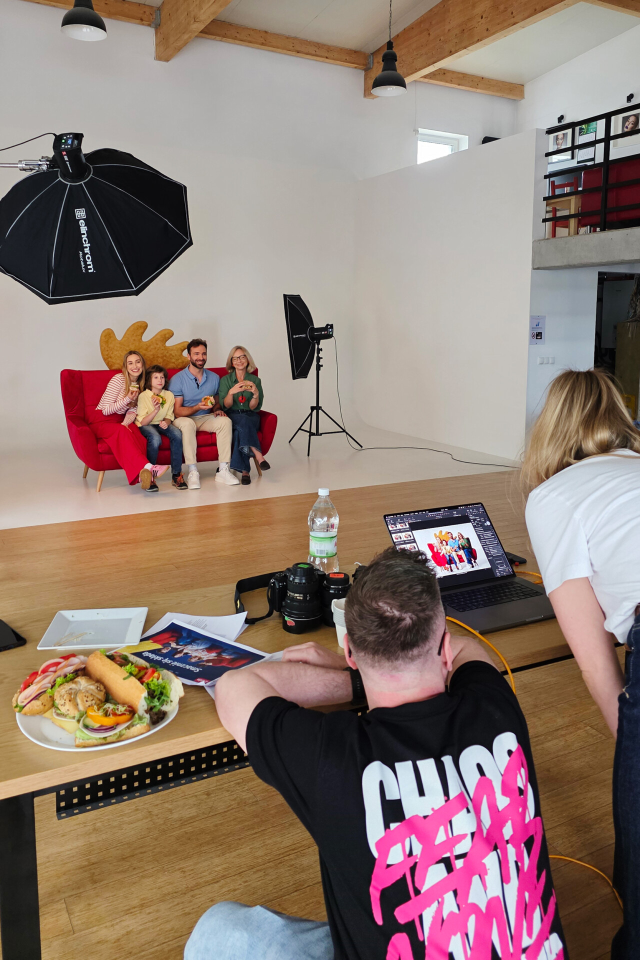 Grupa osób pozujących na czerwonej kanapie przed tłem w studio fotograficznym. W tle widoczny jest sprzęt fotograficzny, a z przodu stół z jedzeniem i laptopem. Jedna osoba zajmuje się ustawianiem sprzętu, podczas gdy reszta pozujących spogląda w obiektyw