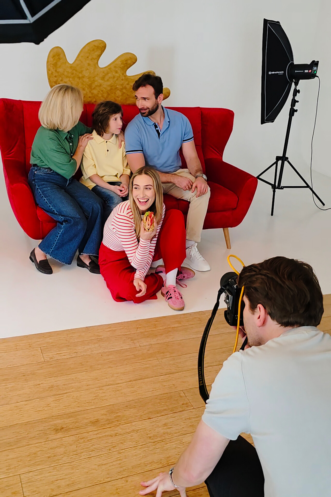 Rodzina pozująca na czerwonej kanapie, z dzieckiem w średnim planie, podczas sesji zdjęciowej. Osoba fotografująca znajduje się na pierwszym planie. W tle znajduje się oświetlenie studyjne i abstrakcyjny kształt