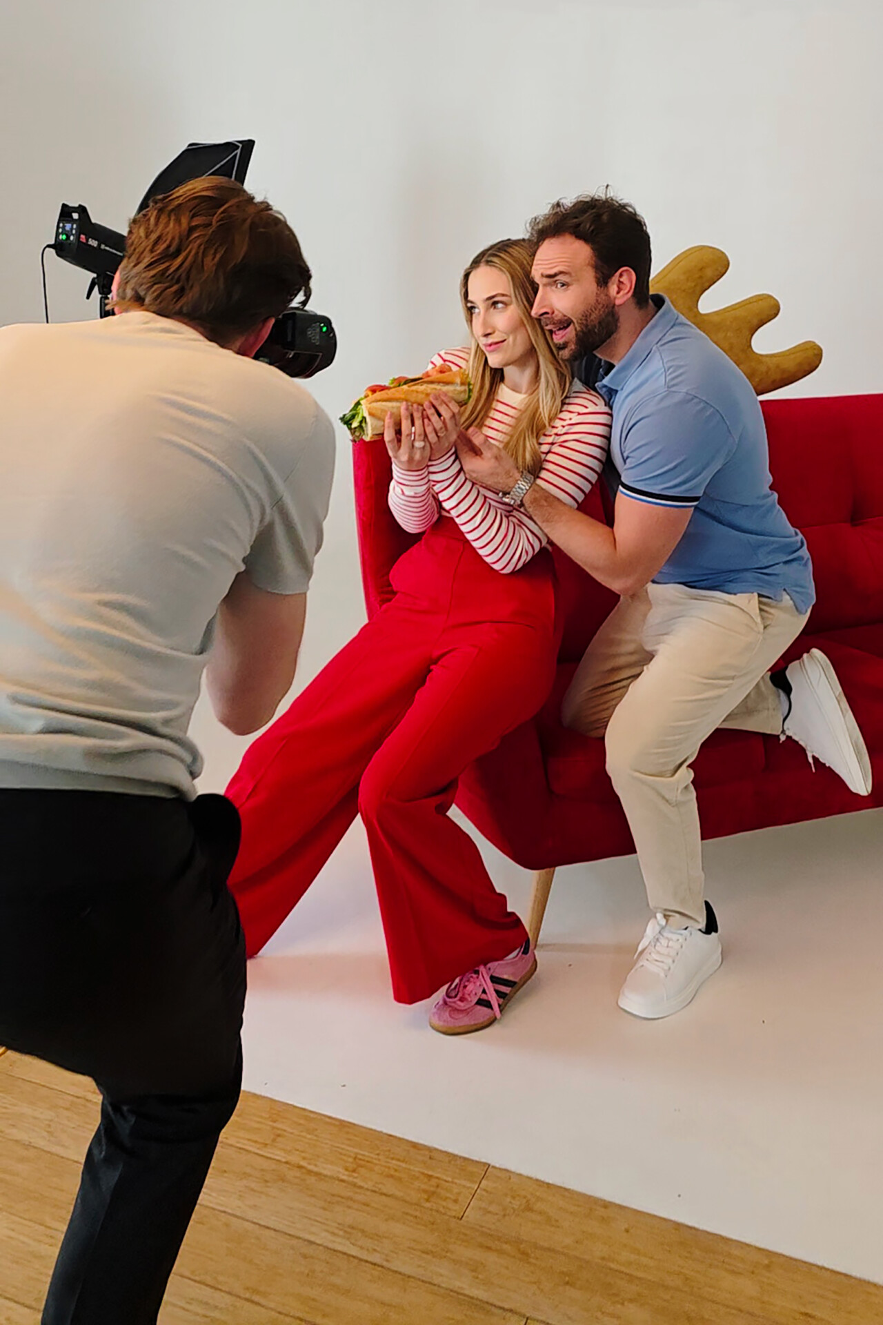 Mężczyzna z kamerą fotografuje parę pozującą na czerwonej kanapie. Kobieta trzyma dużą kanapkę, a mężczyzna obejmuje ją od tyłu, oboje uśmiechają się. W tle znajduje się neutralna, jasna ściana