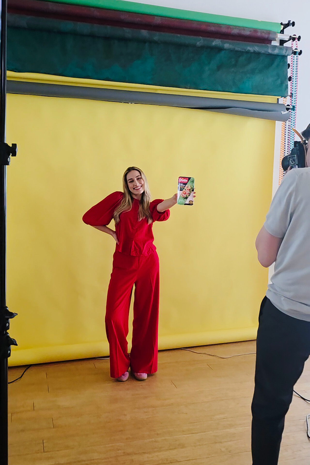 Uśmiechnięta kobieta w czerwonym komplecie modowym trzyma w dłoni ulotkę, pozując na tle żółtej ściany. Obok niej stoi osoba z aparatem fotograficznym