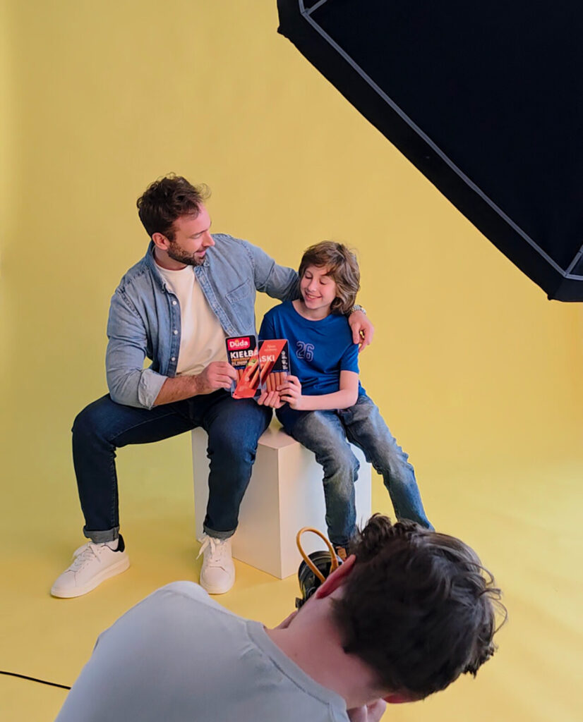 Mężczyzna w jasnej koszuli i dżinsach siedzi obok chłopca w niebieskiej koszulce, obaj trzymają produkt w dłoniach. Tło jest żółte, a ich twarze wyrażają radość. W dolnej części obrazu widoczna jest sylwetka innej osoby, prawdopodobnie fotografa