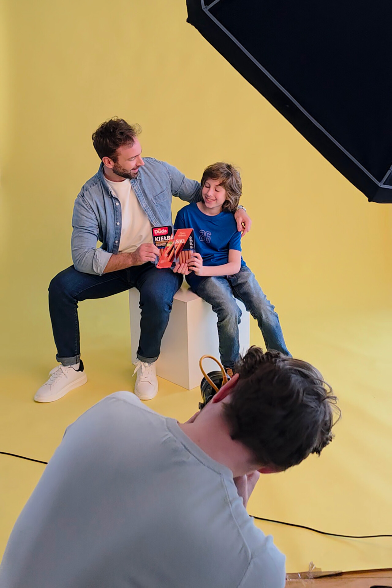 Mężczyzna siedzi na białym sześcianie, trzymając w rękach produkt, obok niego stoi chłopiec w niebieskiej koszulce. W tle jasnoniebieskie tło. Fotografuje ich osoba leżąca na podłodze