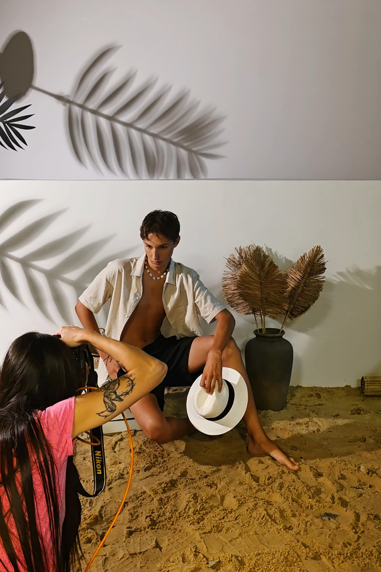 Model siedzący na piasku trzyma kapelusz, podczas sesji zdjęciowej. Fotograf, z długimi włosami i tatuażem, robi zdjęcie, a w tle widać rośliny i dekoracje
