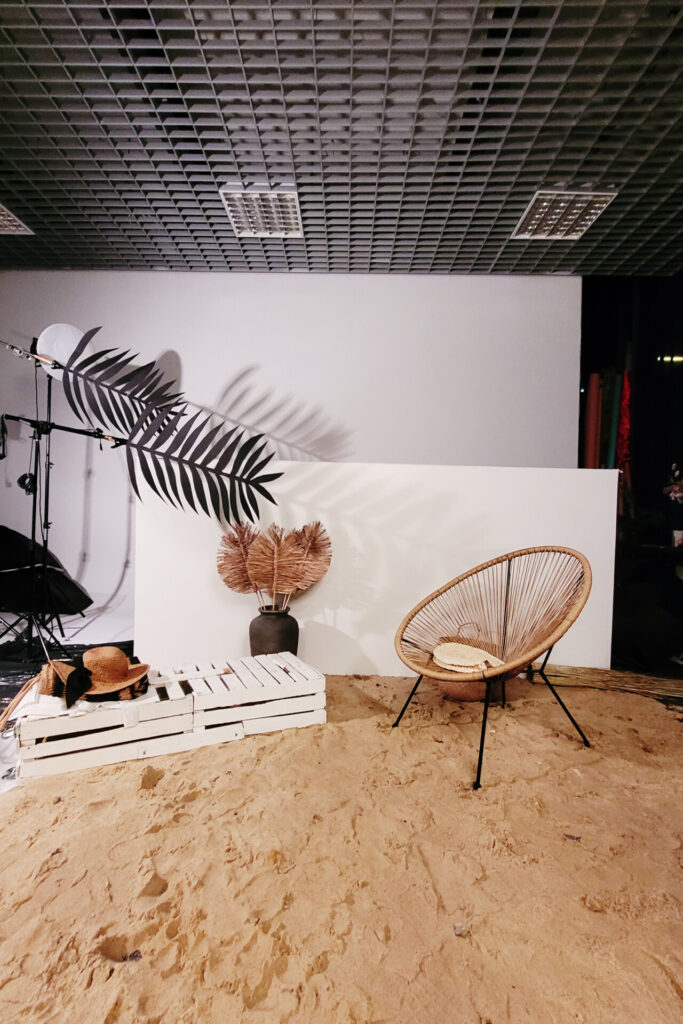 Scena zdjęciowa z piaskiem na podłodze, białą ścianą i dużą palmową gałęzią w tle. Na piasku znajduje się pleciony fotel, a obok niego dekoracyjne elementy, takie jak ciemny wazon z roślinami