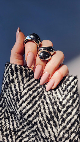 Dłoń z długimi, różowymi paznokciami trzyma dwie srebrne pierścionki, na tle szaro-niebieskiego materiału i wzorzystej tkaniny