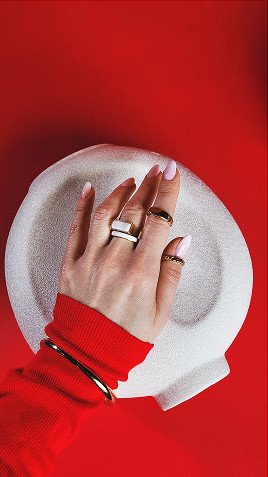 Dłoń z ozdobnymi pierścionkami opiera się na białym naczyniu, na tle intensywnego czerwonego koloru