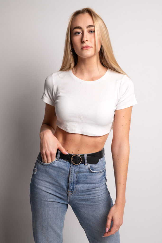 Kobieta w białej, krótkiej koszulce i niebieskich dżinsach z wysokim stanem pozująca na białym tle. Ma długie, proste blond włosy i pewny wyraz twarzy
