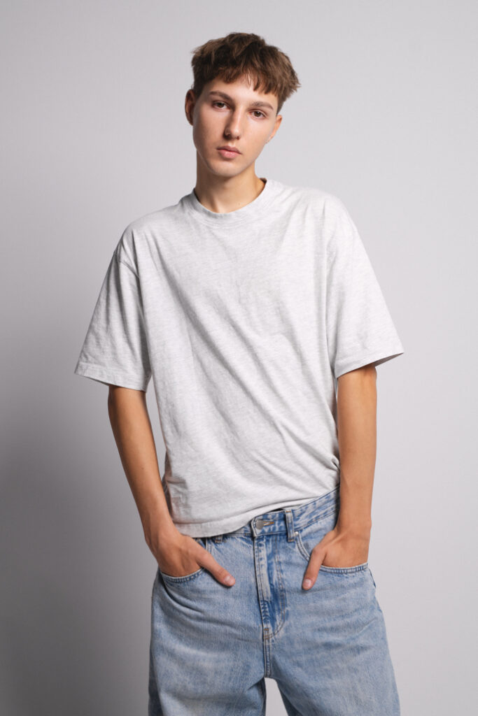 Młody mężczyzna w szarym t-shircie i jasnych, luźnych dżinsach, stoi na tle szarej ściany, z rękami wsuniętymi w kieszenie