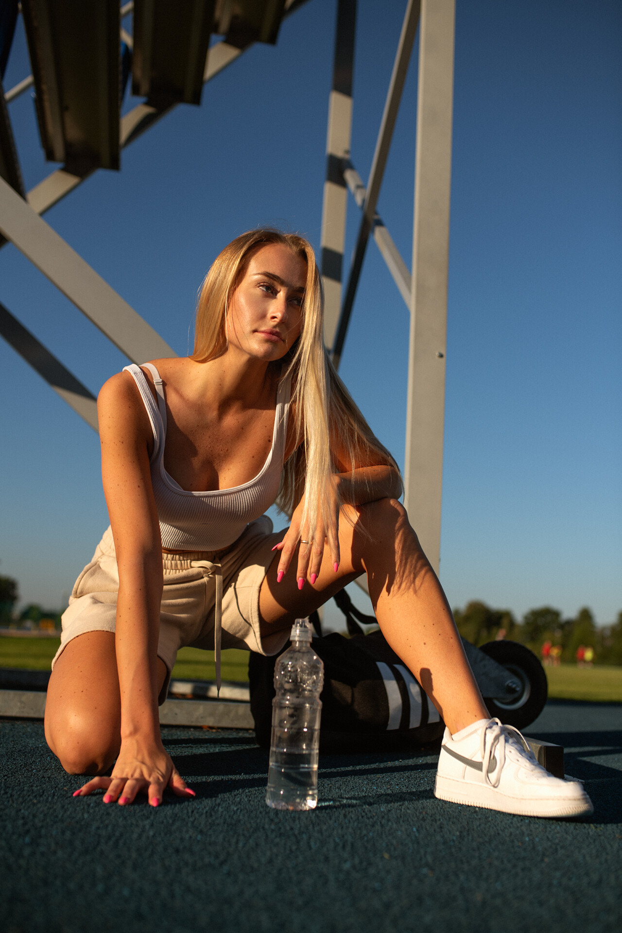 Młoda kobieta w sportowym stroju klęczy na boisku, trzymając rękę na ziemi. Obok niej stoi butelka wody. Tło to zielona przestrzeń z boiskami sportowymi i jasnym niebem