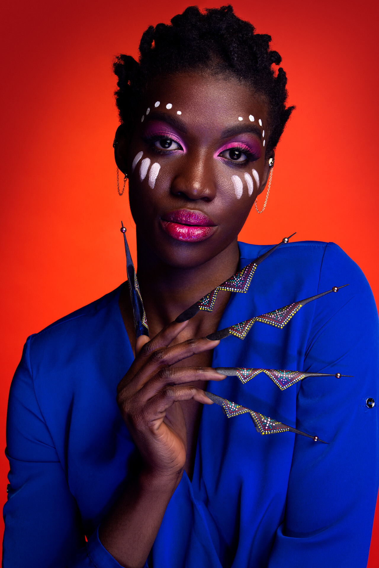 Modelka z długimi, ozdobnymi paznokciami i wyrazistym makijażem, w niebieskiej bluzce, na tle pomarańczowym