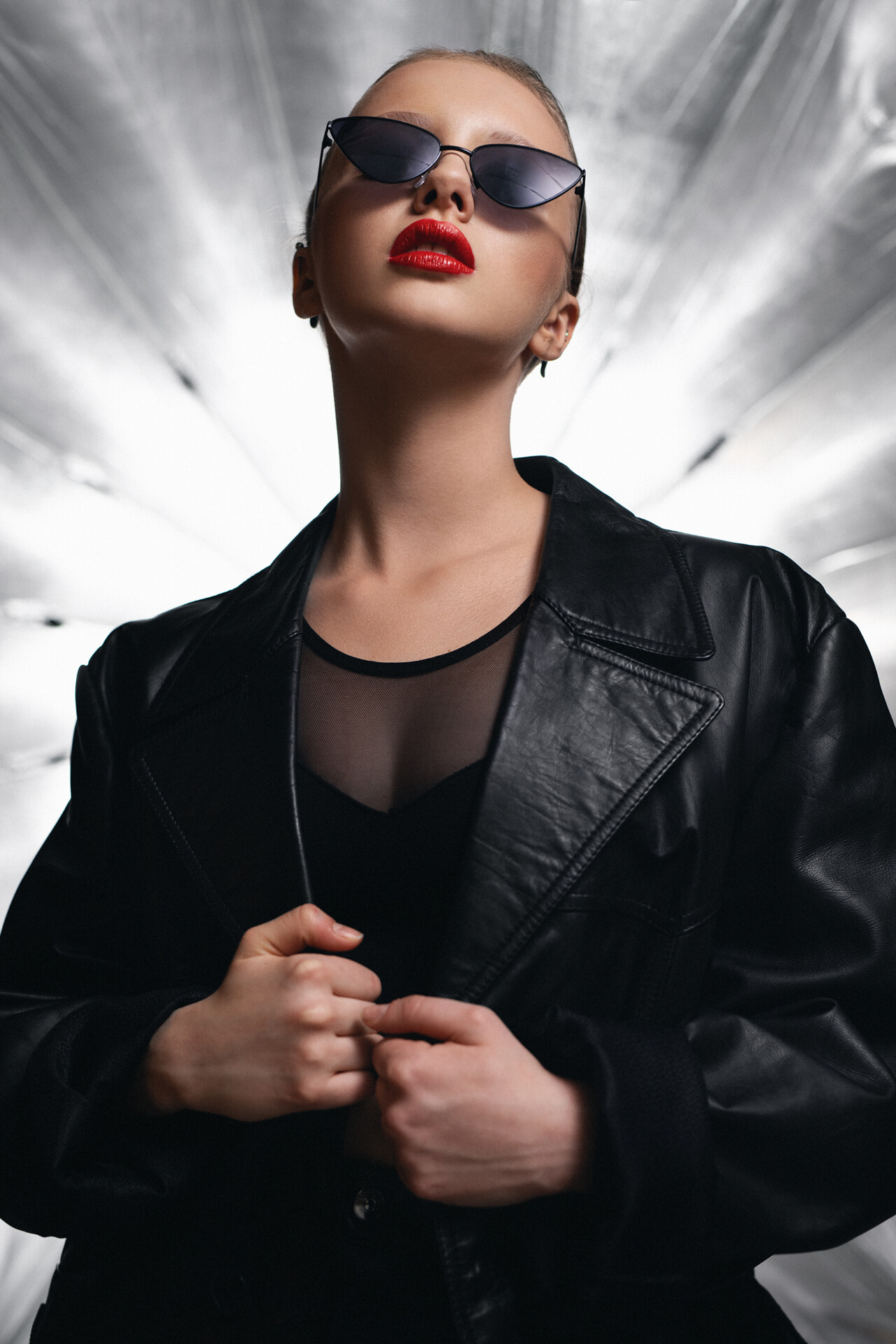 Kobieta w ciemnym, skórzanym płaszczu i okularach przeciwsłonecznych, z czerwoną pomadką, pozuje na tle jasnego, promieniującego tła