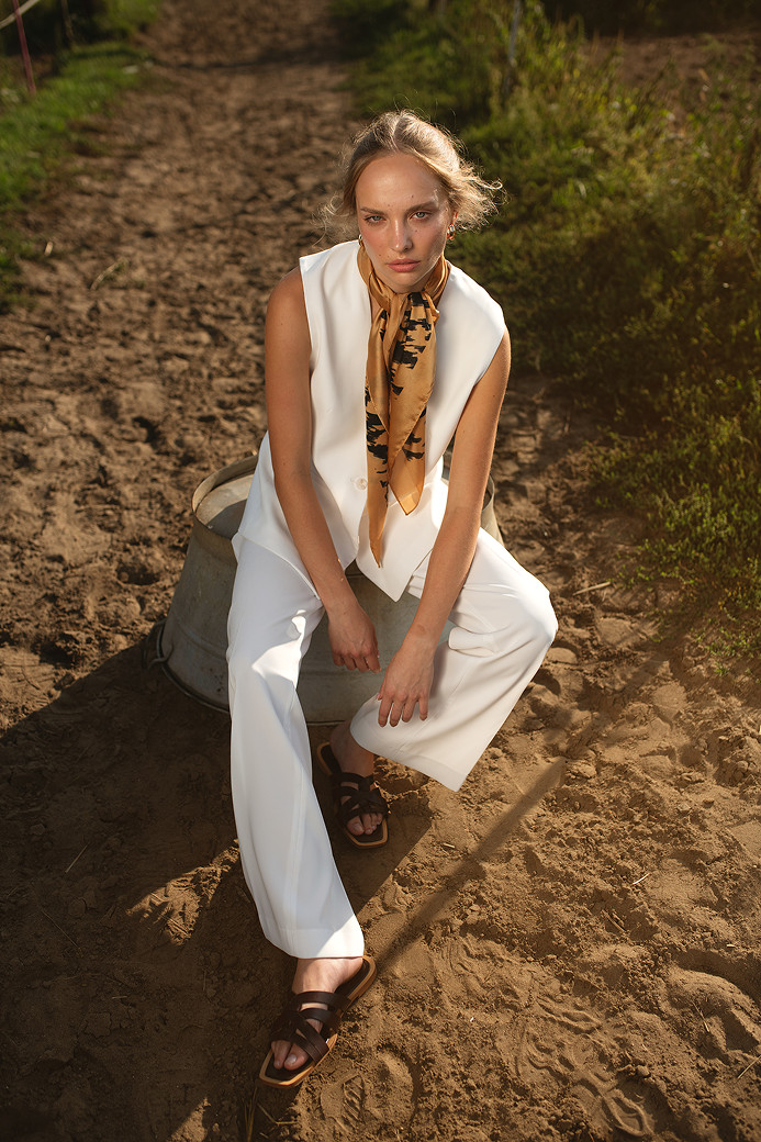 Kobieta w białym kostiumie siedzi na brzegu metalowej beczki, w tle widoczna ziemista ścieżka i zieleń. Ma na sobie kolorowy szal i brązowe sandały, patrzy w stronę obiektywu