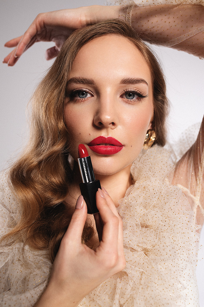 Kobieta z falującymi włosami trzyma w dłoni czerwony błyszczyk do ust, patrząc bezpośrednio w obiektyw. Ubrana w jasną, elegancką sukienkę z dużymi rękawami, tło jest stonowane i jasne