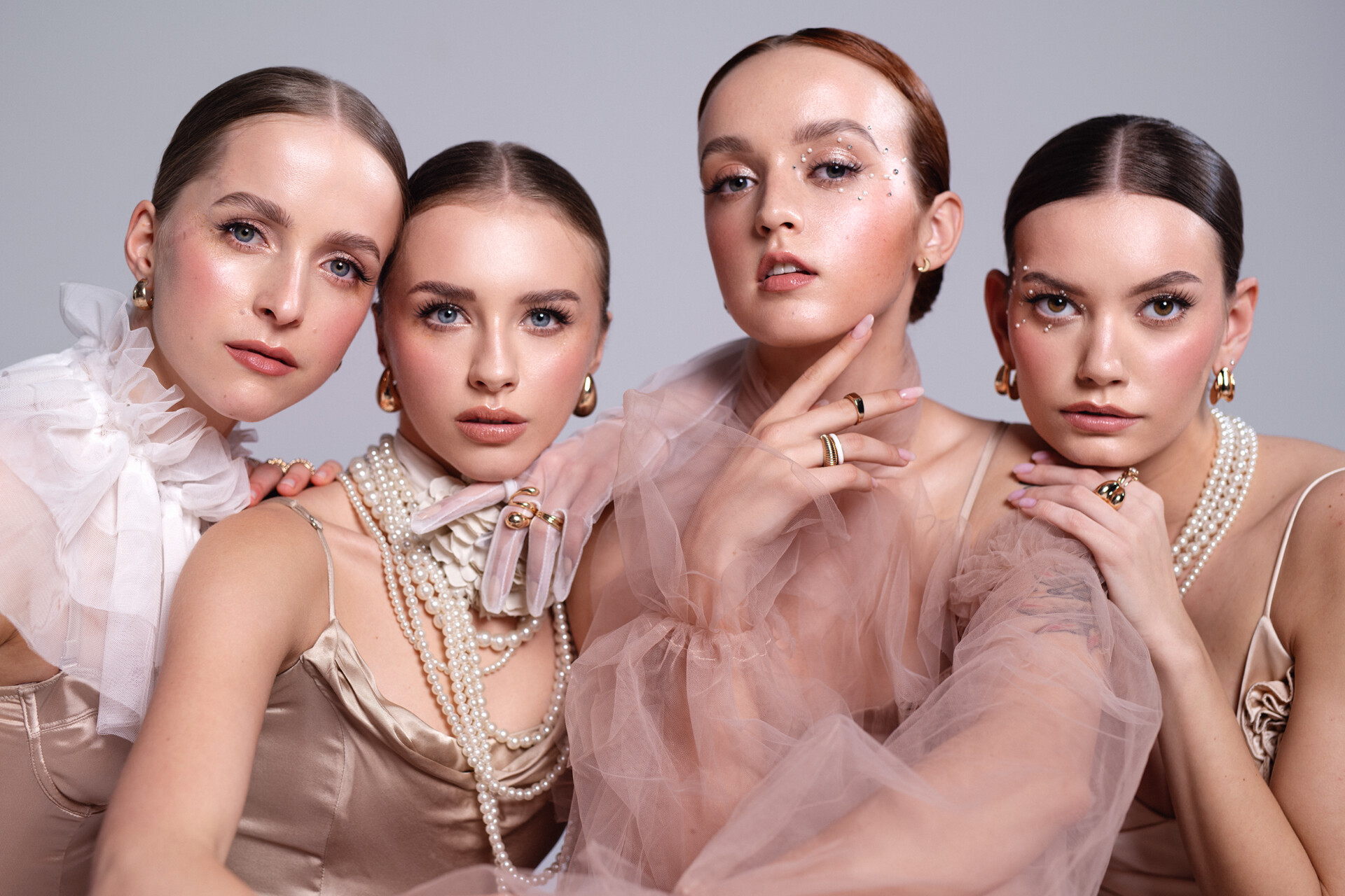 Cztery kobiety w eleganckich strojach w kolorze nude, siedzą obok siebie. Każda z nich ma stylizowane fryzury i ozdoby, w tym biżuterię oraz perły. Użyto delikatnych makijaży, a ich wyraziste spojrzenia skierowane są w stronę obiektywu. Tło jest w odcieniach szarości