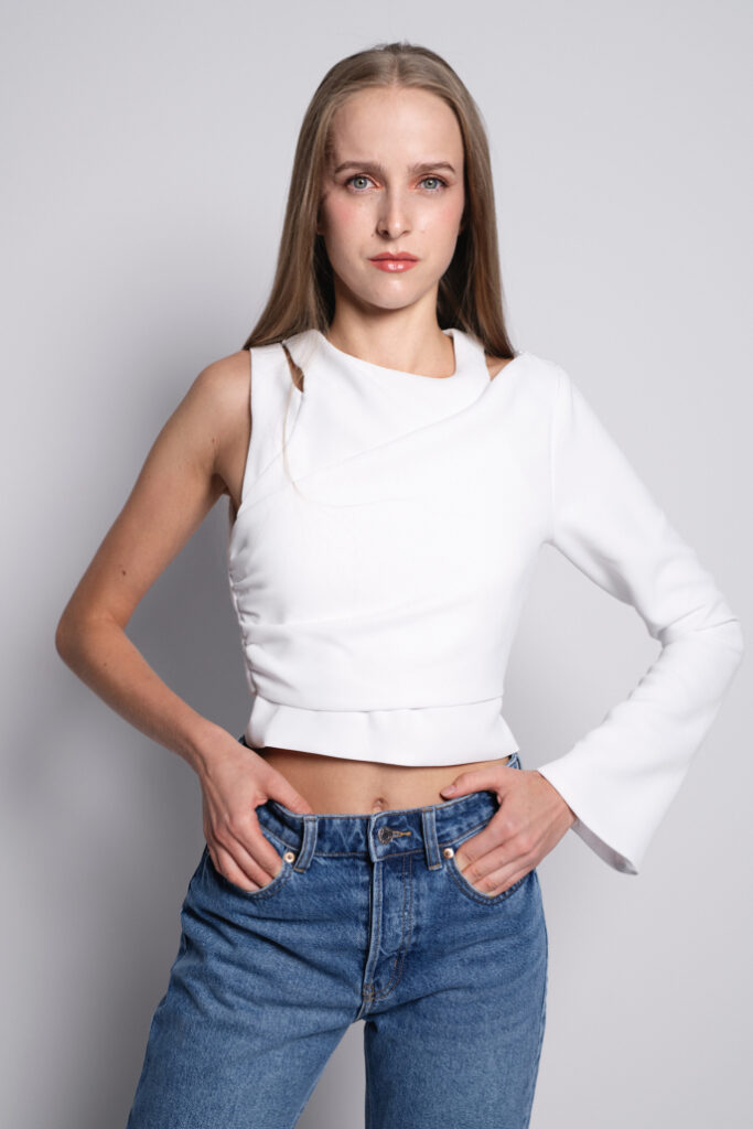 Kobieta w białej bluzce o asymetrycznym kroju, z długim rękawem z jednej strony i krótkim z drugiej, stoi w lekkim profilu, ręce na biodrach, z uśmiechem na twarzy, w obcisłych niebieskich dżinsach. Tło jest jasnoszare