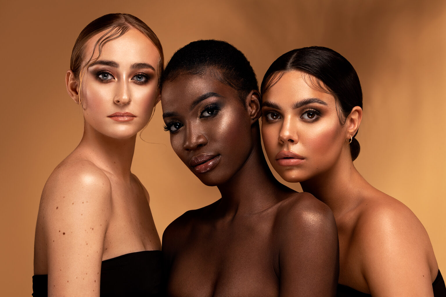Trzy kobiety o różnym kolorze skóry stoją blisko siebie, każda z delikatnym makijażem i eleganckimi fryzurami. Tło jest w ciepłych odcieniach brązu, co podkreśla ich rysy