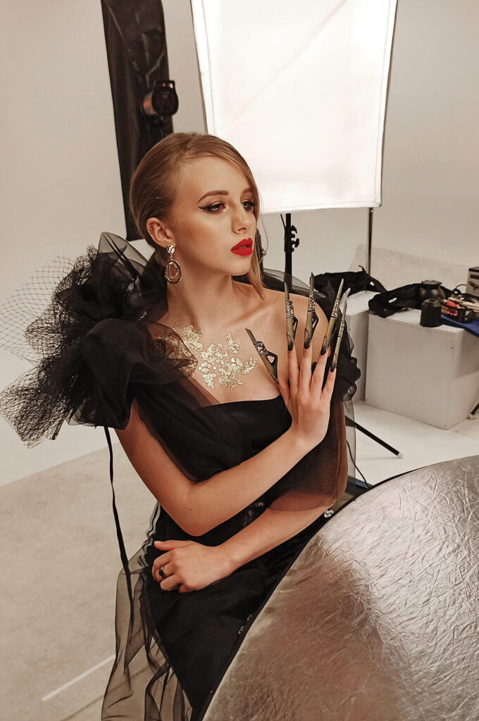 Kobieta w czarnej sukience z ozdobnymi rękawami, z dramatycznym makijażem i długimi, stylowymi paznokciami, siedzi przy stole w studiu. W tle widoczne oświetlenie studyjne i sprzęt fotograficzny