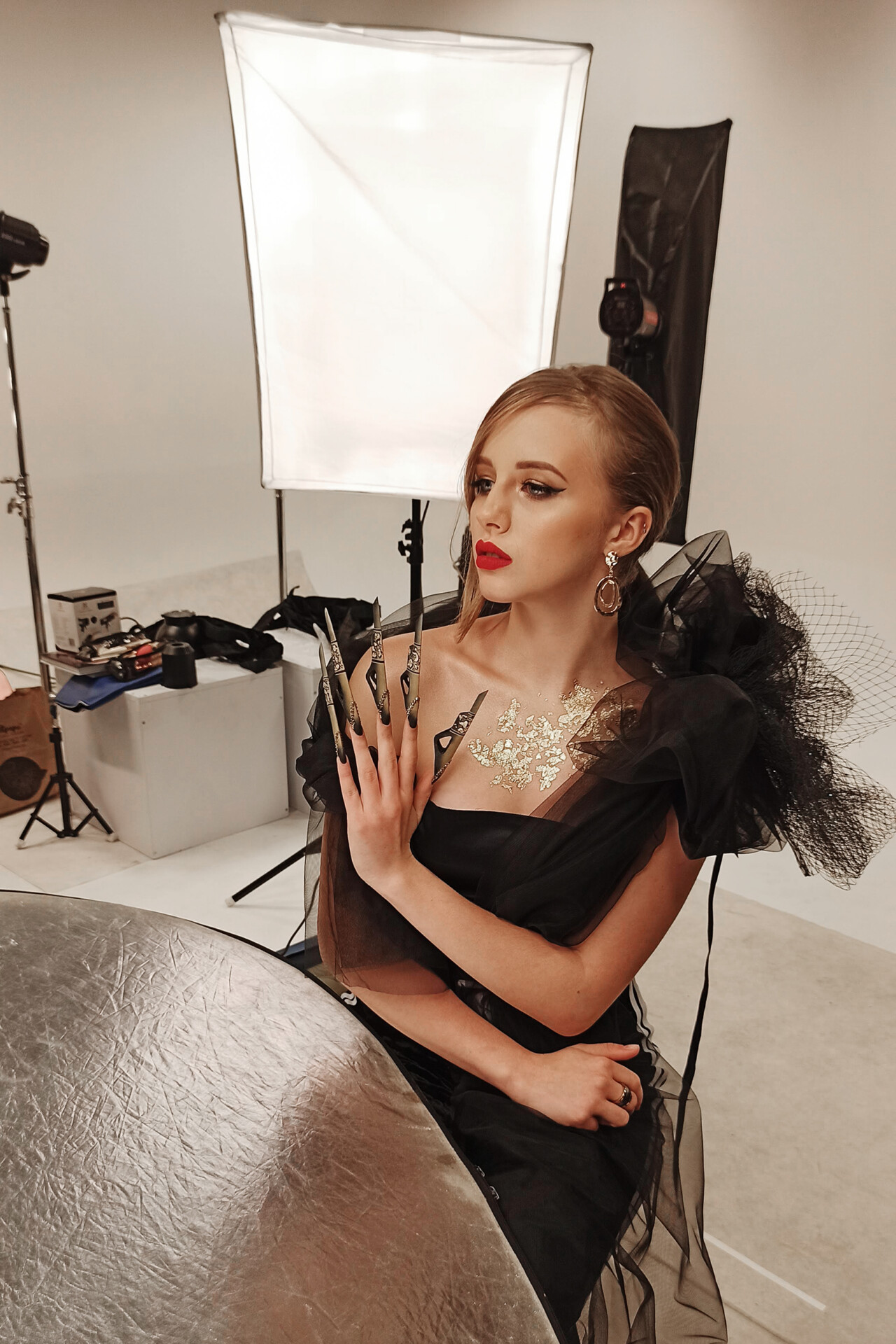 Kobieta w eleganckiej czarnej sukni z dużymi rękawami z tiulu, trzymająca ozdobne paznokcie, siedzi przy okrągłym stole. W tle widoczna scena fotograficzna z lampami