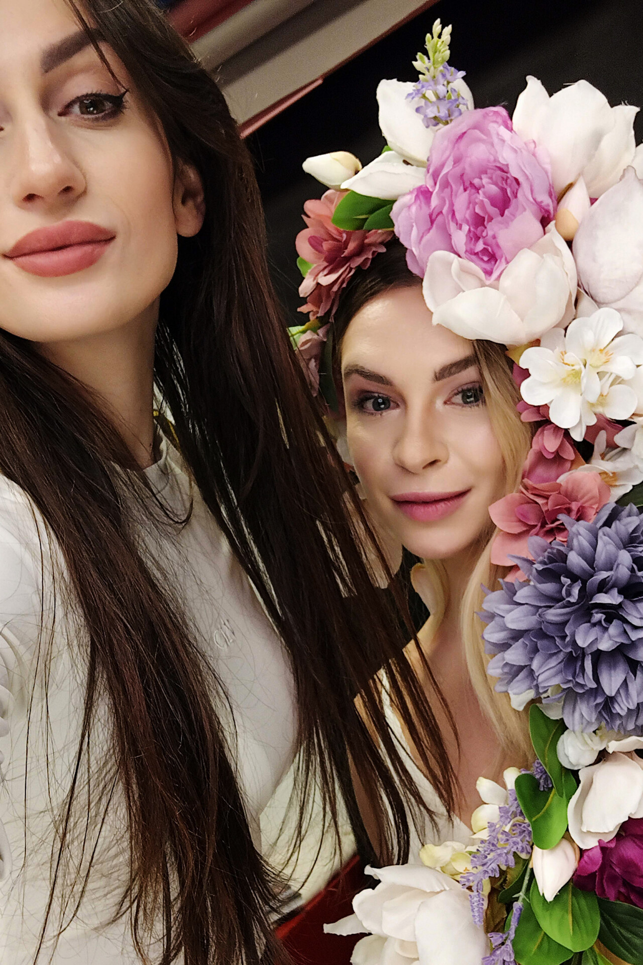Dwie kobiety pozują do selfie, jedna z nich ma na sobie kwiecisty wianek w pastelowych kolorach, a obie uśmiechają się do kamery
