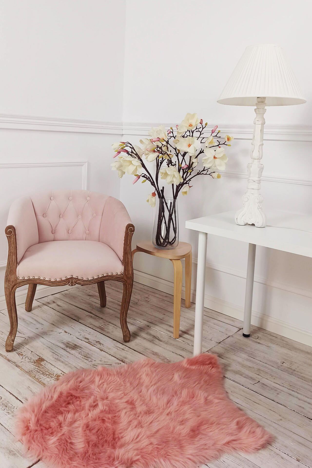 Różowe, welurowe krzesło obok eleganckiego stolika w jasnym kolorze, na którym stoi lampa oraz wazon z kwiatami. Na podłodze leży różowy, futrzany dywan