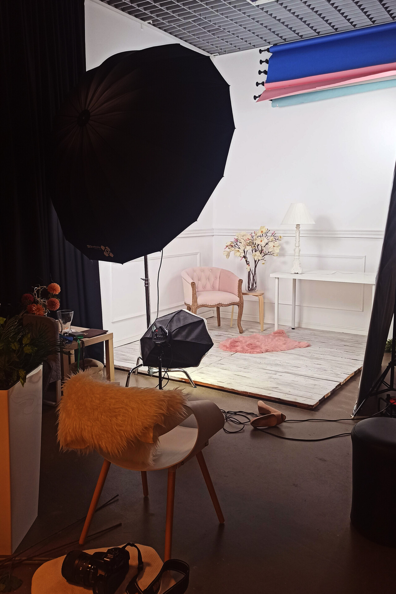 Studio fotograficzne z różowym fotelem, białym stół i dekoracyjnymi elementami, na podłodze dywanik, oświetlenie w tle