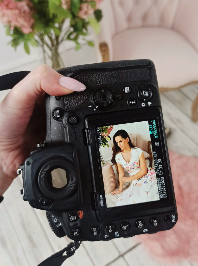 Kamera trzymana w dłoni, na wyświetlaczu widoczne zdjęcie kobiety w kolorowej sukience siedzącej na różowej sofie; w tle widać kwiaty