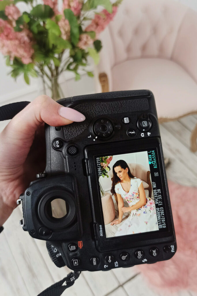 Aparat fotograficzny trzymany w dłoni, na ekranie wyświetlana jest kobieta w kwiecistej sukience, siedząca na różowym fotelu, otoczona kwiatami