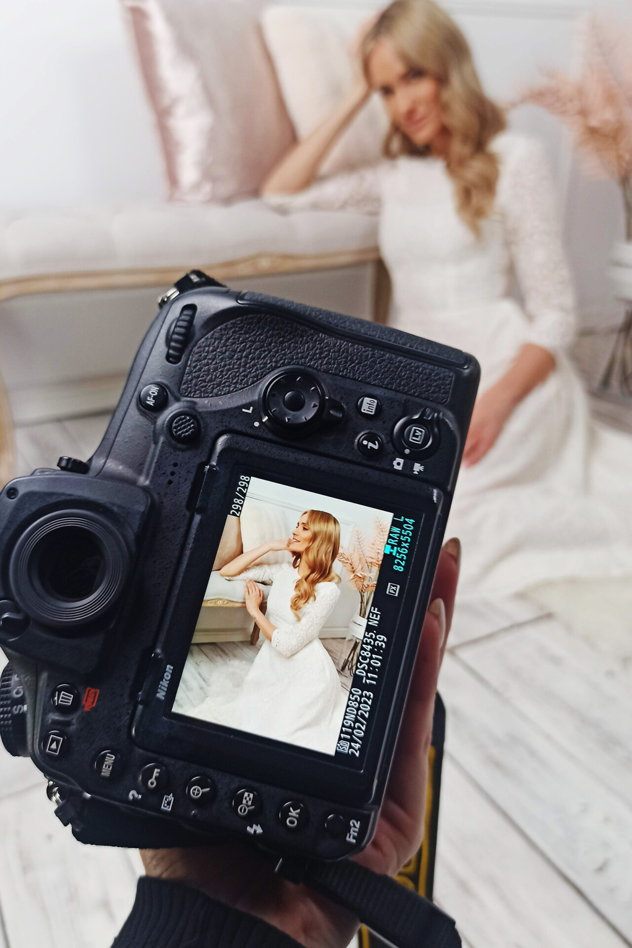 Kobieta w jasnej sukni siedzi na kanapie, a przed nią widoczny jest aparat fotograficzny z wyświetlaczem pokazującym jej ujęcie. W tle delikatne dekoracje