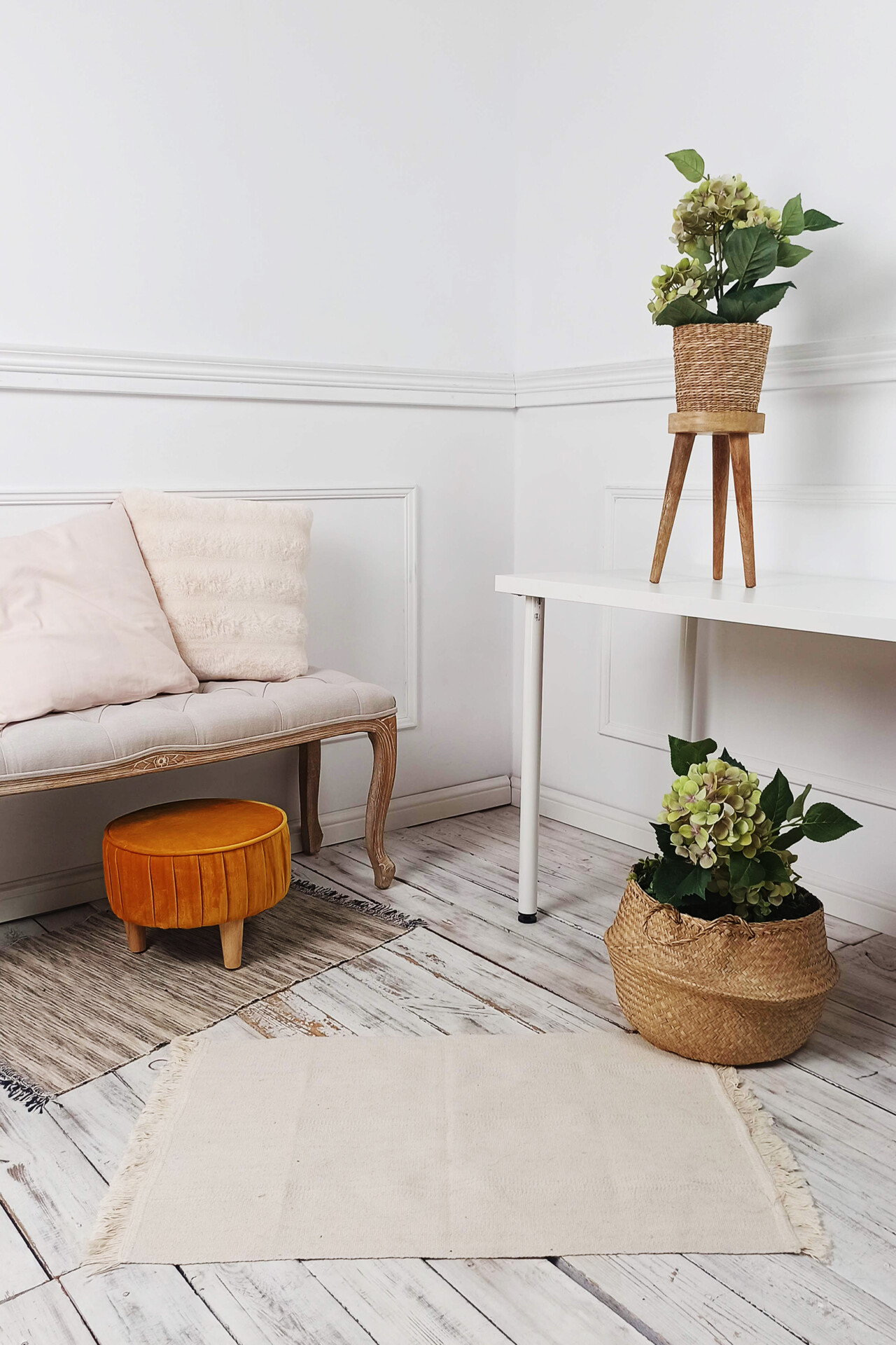 Nowoczesny kącik wnętrza z jasną podłogą, sofą z jasnymi poduszkami, niskim, okrągłym stolikiem oraz roślinami w koszach. W tle widać biały stół z nogami w stylu skandynawskim