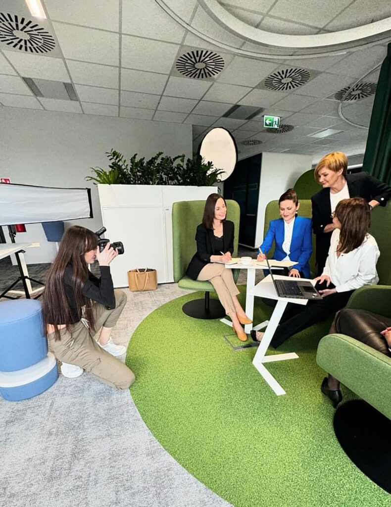 Grupa kobiet siedzi przy stole w biurze, podczas sesji zdjęciowej. Jedna z kobiet robi zdjęcia, pozostałe oglądają dokumenty i pracują na laptopie. Otoczenie jest nowoczesne, z zielonym dywanem i przestronnymi elementami dekoracyjnymi