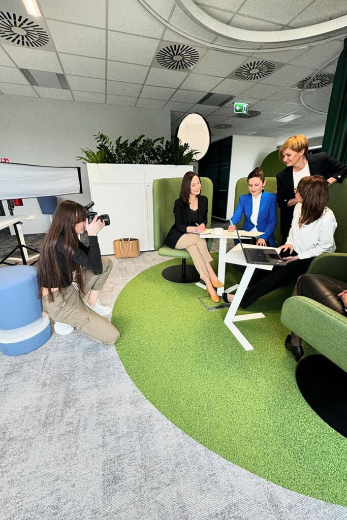 Grupa pięciu kobiet w formalnych strojach siedzi przy stole w nowoczesnym biurze, omawiając dokumenty. Jedna z kobiet fotografuje scenę, a dwie inne angażują się w rozmowę. W tle widoczne zielone elementy dekoracyjne i funkcjonalne meble biurowe