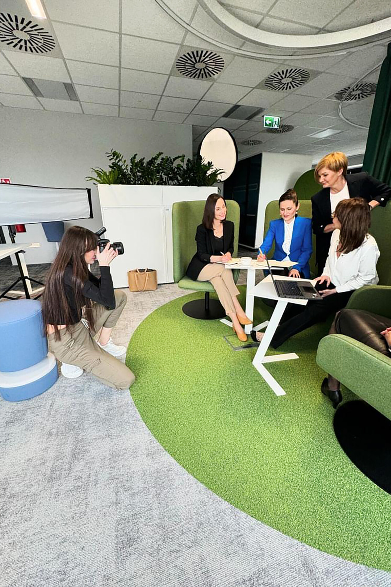 Grupa pięciu kobiet w formalnych strojach siedzi przy stole w nowoczesnym biurze, omawiając dokumenty. Jedna z kobiet fotografuje scenę, a dwie inne angażują się w rozmowę. W tle widoczne zielone elementy dekoracyjne i funkcjonalne meble biurowe